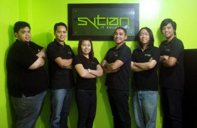 Six members of the Sytian IT Solutions team stand side-by-side in matching black polo shirts in front of a lime green wall with the company sign hanging behind them.