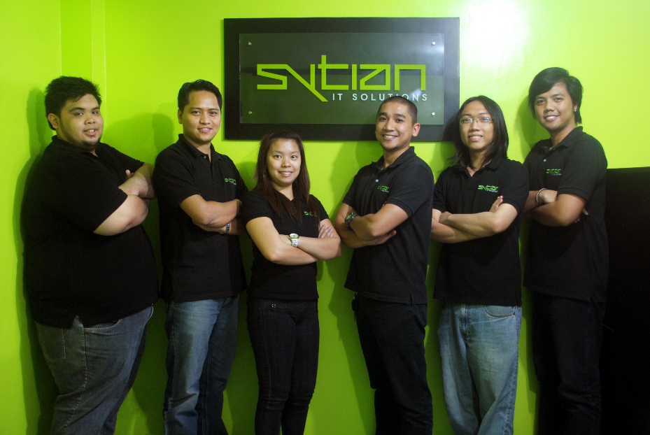 Six members of the Sytian IT Solutions team stand side-by-side in matching black polo shirts in front of a lime green wall with the company sign hanging behind them.