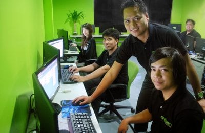 Four young adults sit at desks in an office with bright green walls, facing computer screens with keyboards and mice, while a smiling man leans over one desk to look at the camera.