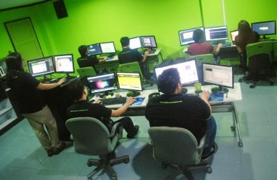 Several people work on computers in a brightly lit office with green walls and grey tiled floors.