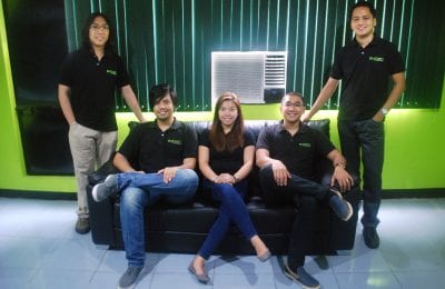 A group of five smiling colleagues, three men and a woman, pose for a photo in an office with bright green walls and a dark leather sofa.