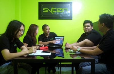 Five employees in black polo shirts collaborate around a table in a bright green office, with a laptop displaying a website and the Sytian IT Solutions logo mounted on the wall behind them.