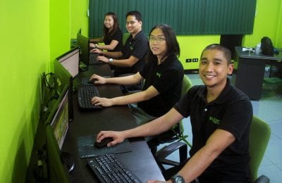 Four people sitting at desks in a row in an office with bright green walls, all smiling and looking towards the camera, with computers in front of them.