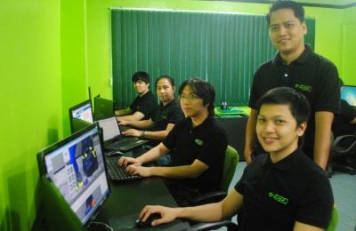 A group of five men wearing black company shirts with green accents working at computer stations in an office with bright green walls.