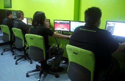 Four employees sit at a row of computers against a lime-green wall, each using a matching black and neon-green office chair.
