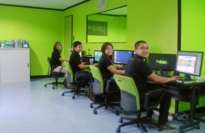 Four young professionals are seated at desks in an office with bright green walls, each working on their own computer and smiling toward the camera.