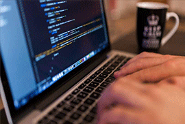 A person typing on a laptop keyboard with code displayed on the screen and a coffee mug in the background.