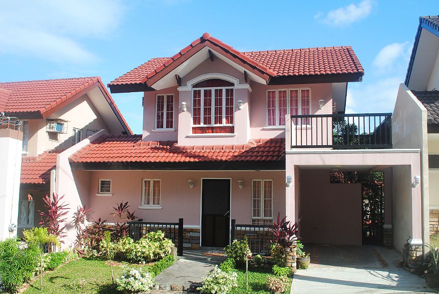 A two-story pink house featuring a red-tiled roof, a balcony, and a covered garage stands prominently under a bright blue sky.
