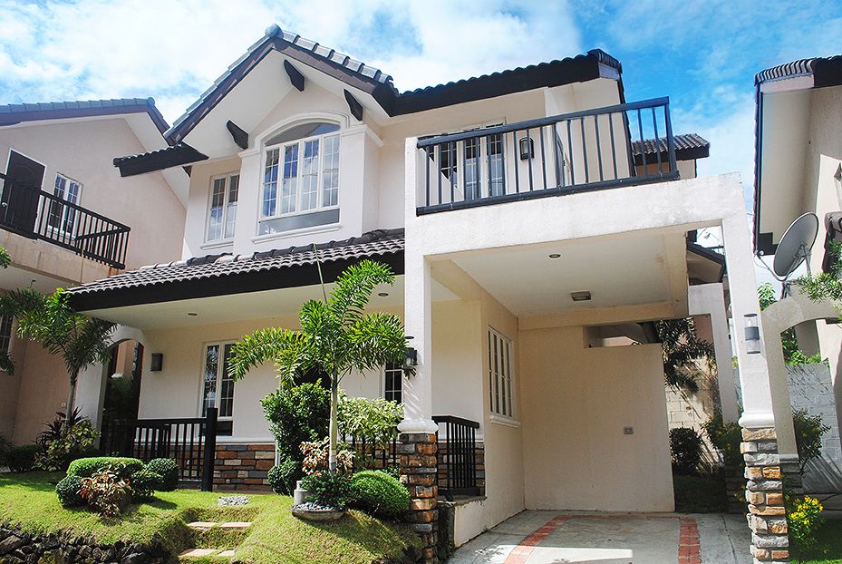 A two-story cream-colored house featuring a black-tiled roof, an upper-level balcony with a black railing, and a covered carport below under a partly cloudy sky.