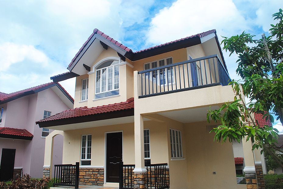 A two-story beige house with a red tile roof features a second-floor balcony and stone accents around the first-floor porch.