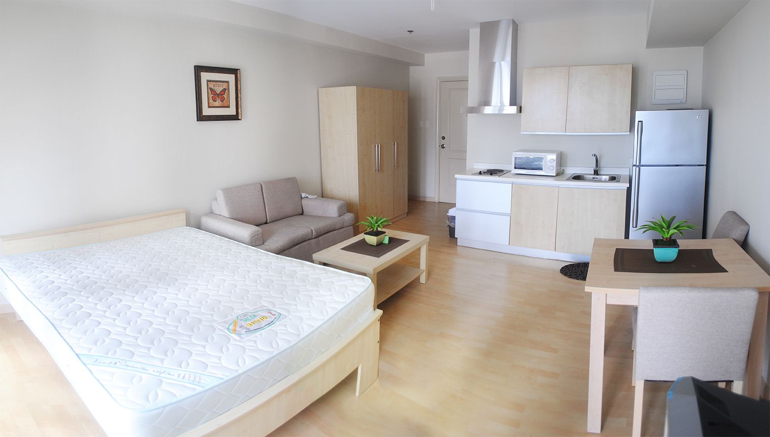Studio apartment interior featuring a bed with a white mattress, a small sofa, a coffee table, a dining table with chairs, a wardrobe, and a compact kitchen area with a refrigerator and stove.