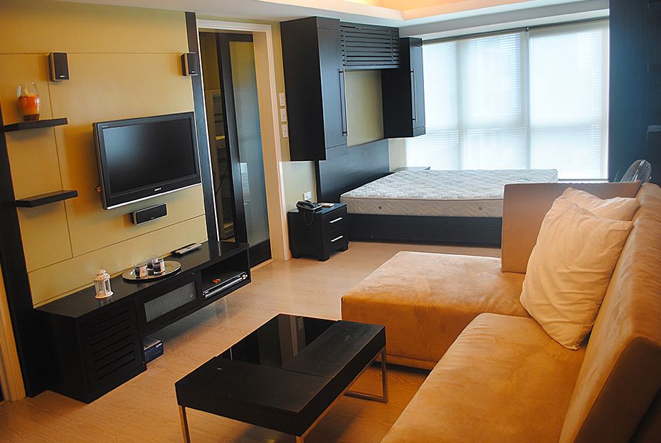 A well-lit modern living room featuring a tan sectional sofa, a black coffee table, a TV mounted on a tan wall, and a bed in the background near a window.