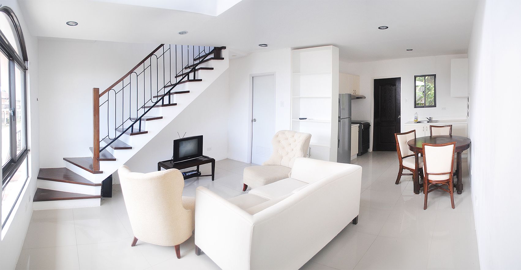 A bright, modern open-plan living room with white walls, tiled floors, and a staircase leading upstairs, furnished with a white sofa, armchairs, a TV, and a dining area in the background.