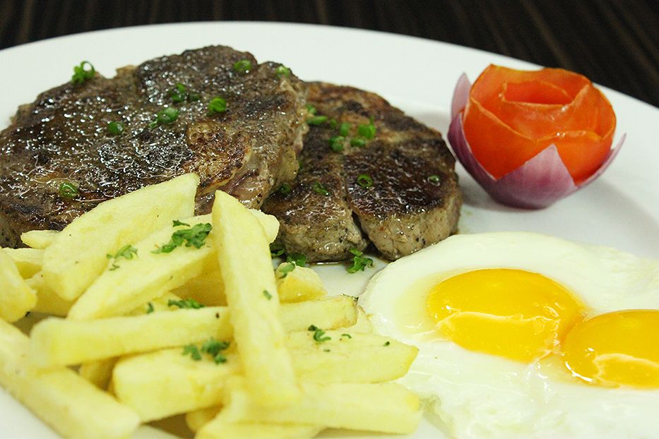 A white plate holds two grilled steaks topped with chives, a pile of thick-cut french fries garnished with herbs, two sunny-side-up eggs, and a tomato carved into a rose shape nested in red onion petals.