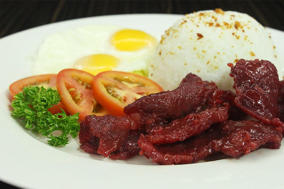 A white plate holds a Filipino breakfast meal with sweet cured beef tapa, garlic fried rice, two sunny-side-up eggs, sliced tomatoes, and fresh parsley garnish.