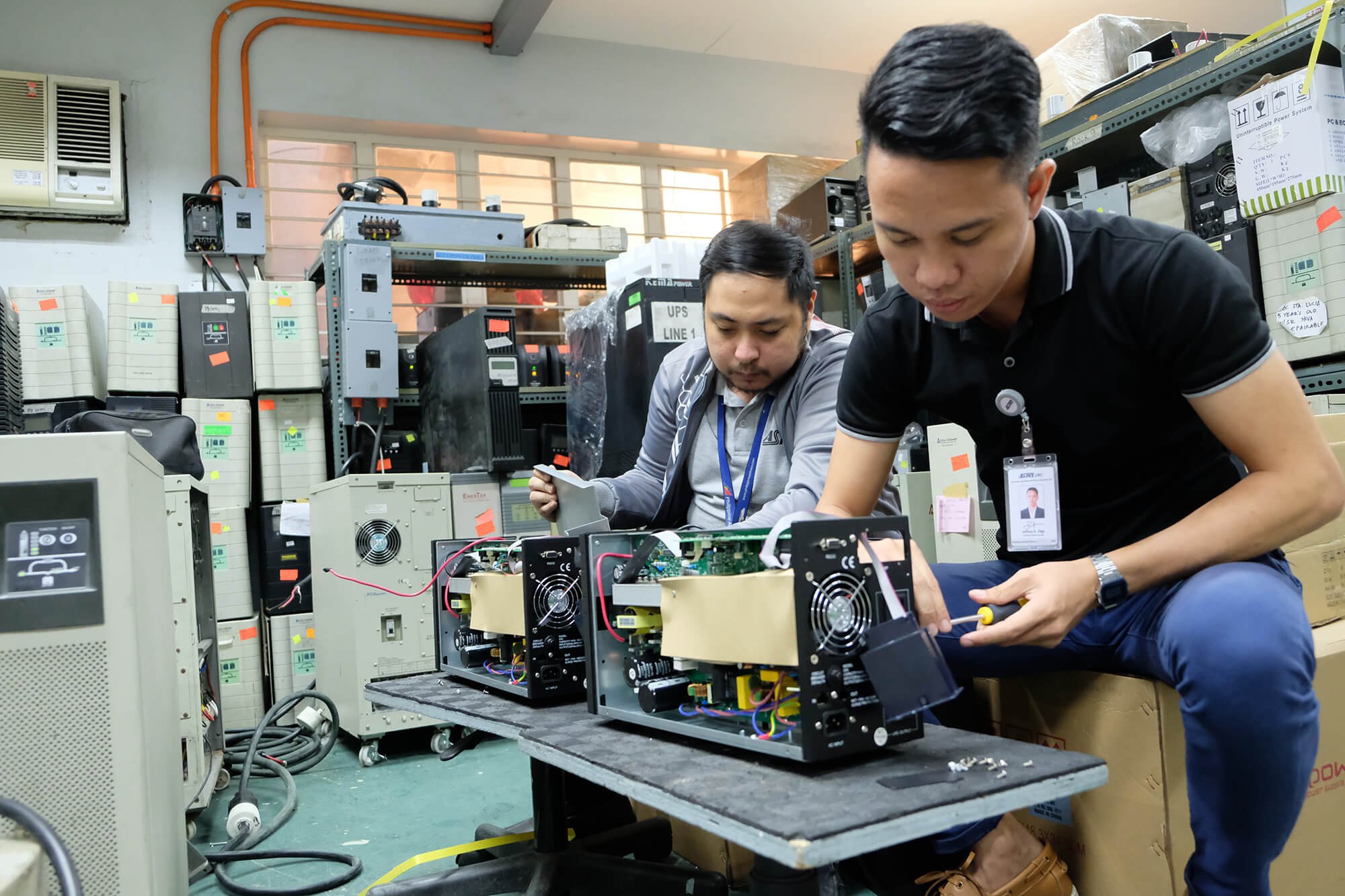 Two men in a workshop repair open electrical devices on a table, surrounded by stacked equipment and storage shelves.