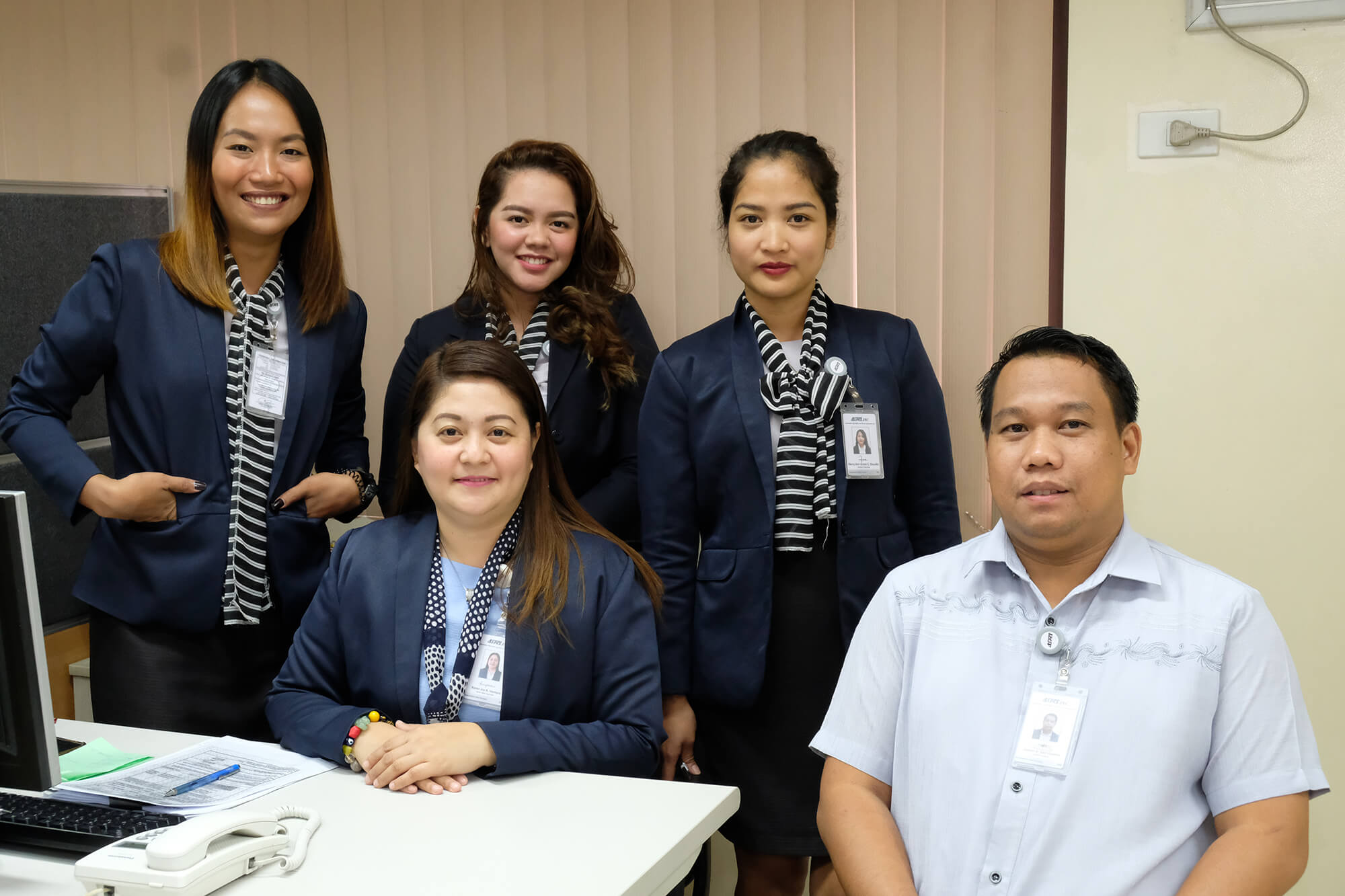 A group of five office workers, including four women in matching navy blue blazers with striped scarves and one man in a light blue button-down shirt, pose together in an office setting.