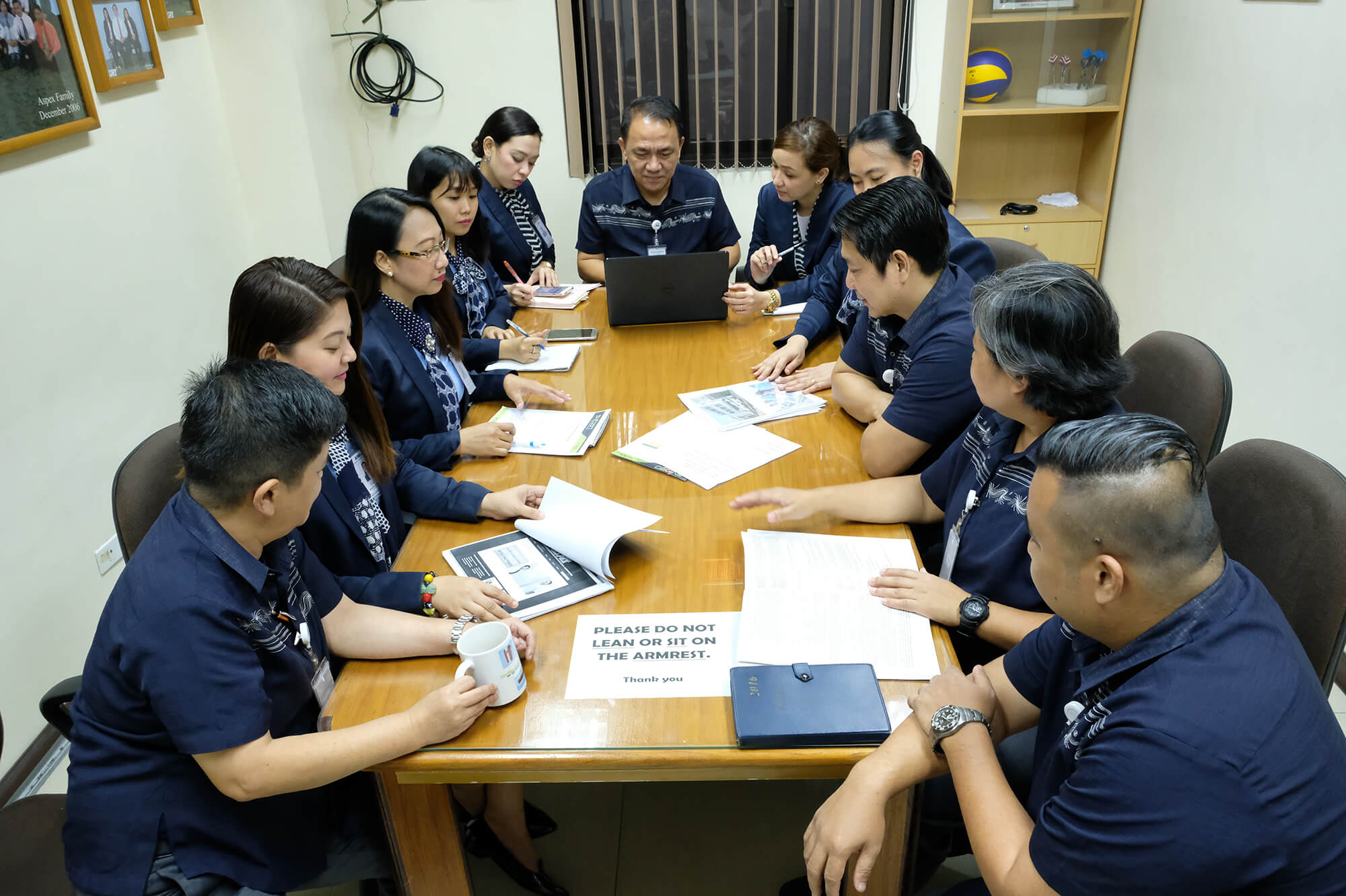 A group of people wearing blue polo shirts sit around a long wooden conference table, reviewing documents and discussing work in a brightly lit office.