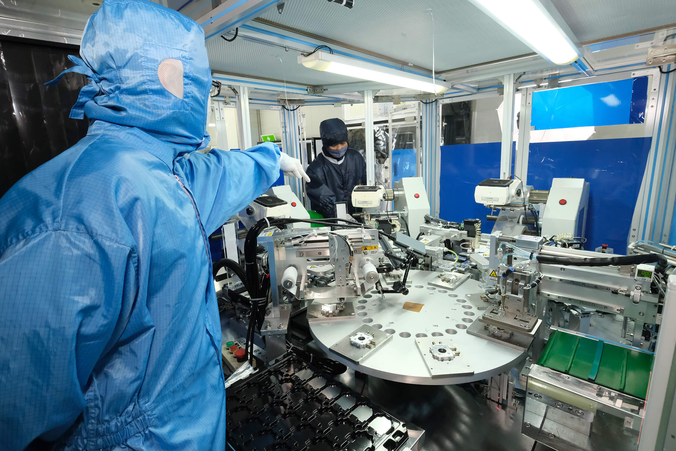 Two workers in cleanroom suits operate industrial machinery in a bright factory setting.