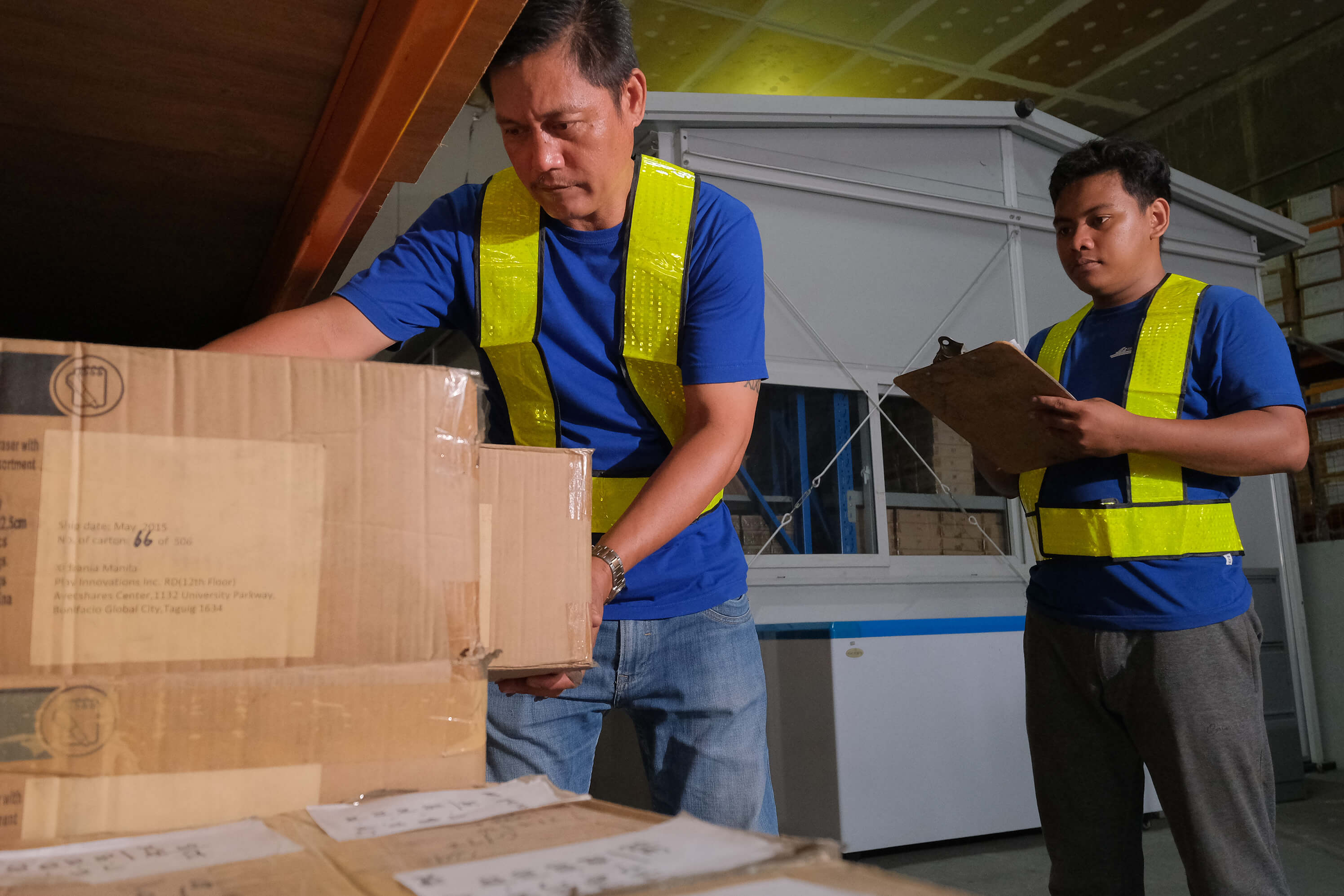 Two warehouse workers in blue shirts and yellow high-vis vests manage inventory, with one lifting cardboard boxes and the other holding a clipboard.