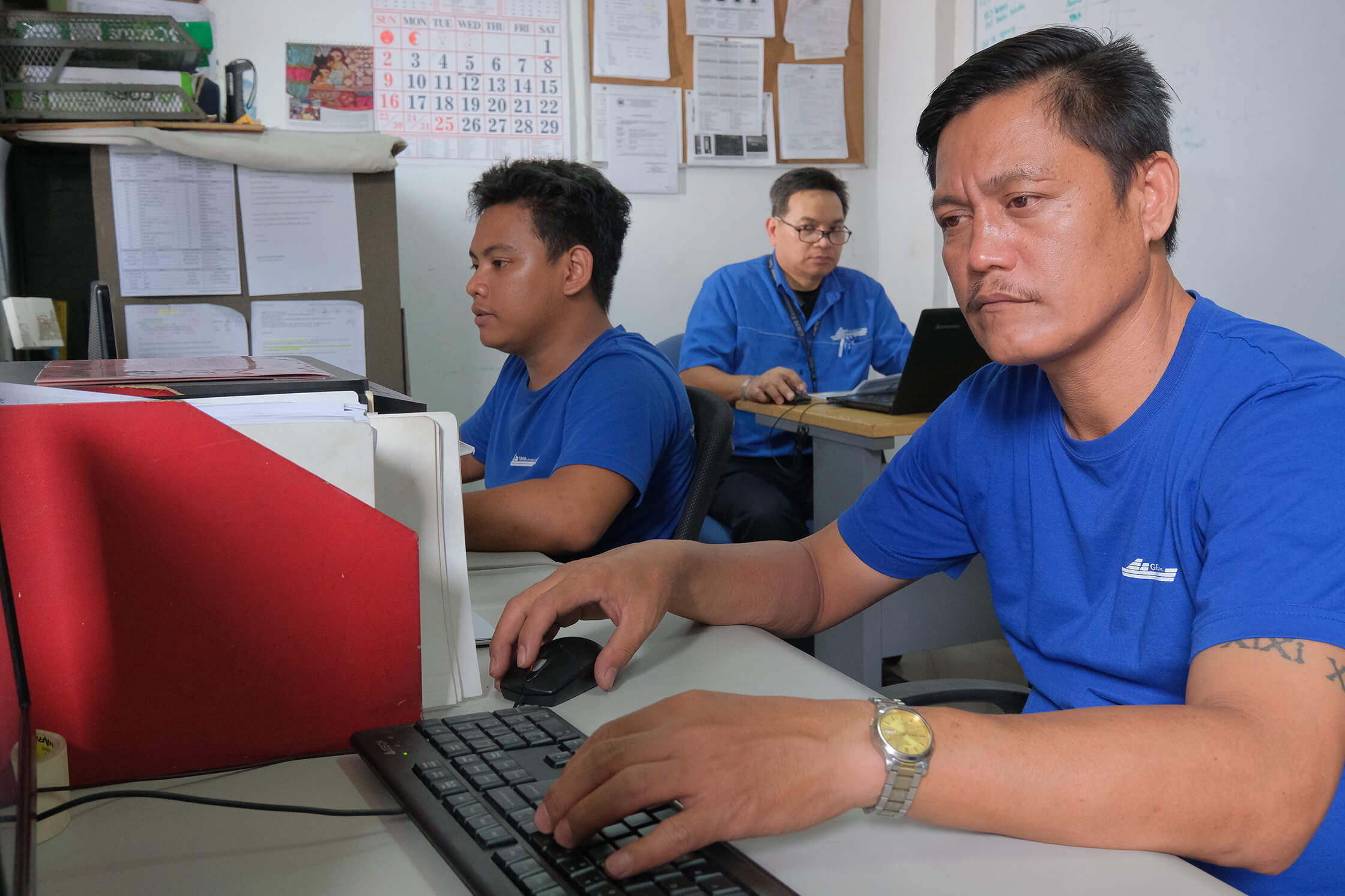 Three men wearing blue shirts work at their computers in an office setting.