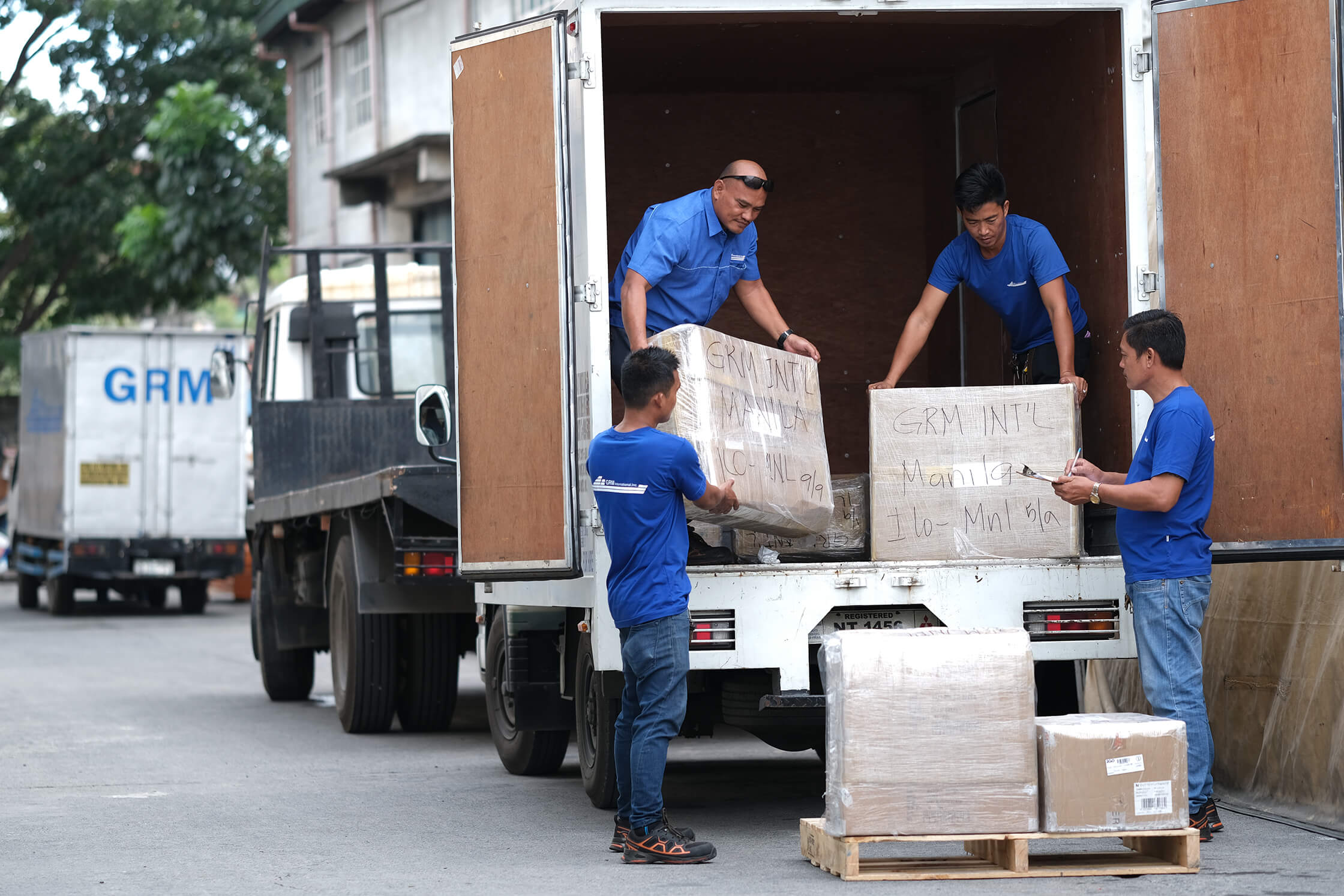 Four men in blue uniforms load large boxes into the back of a delivery truck, with another truck parked behind it.