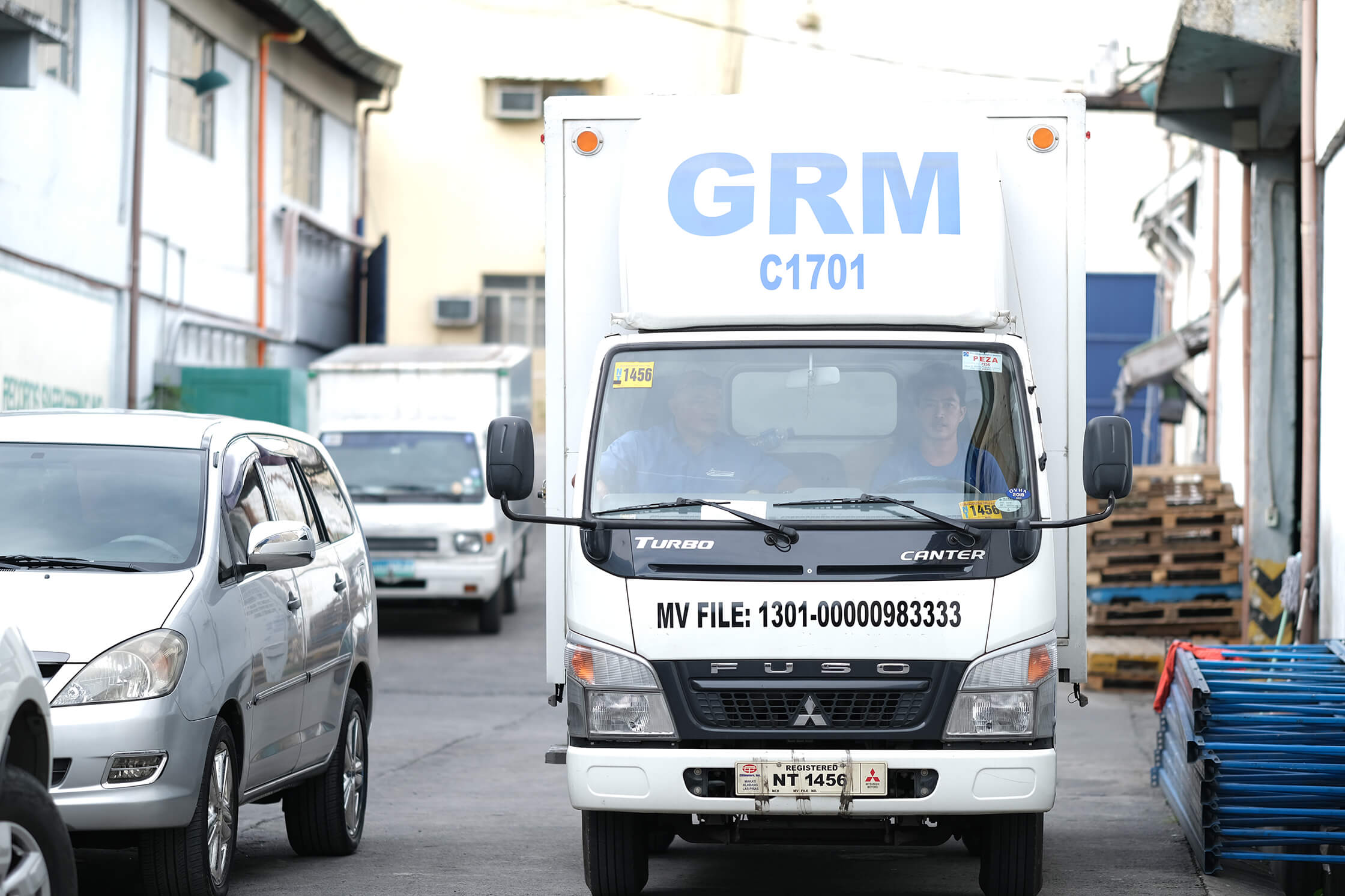 A white GRM delivery truck approaches on a paved industrial street, flanked by a silver car on the left and wooden pallets on the right.