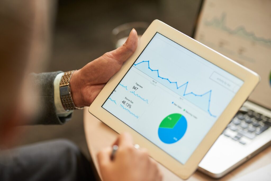 A person holding a tablet computer displaying a financial line chart and pie chart, with a laptop in the background.