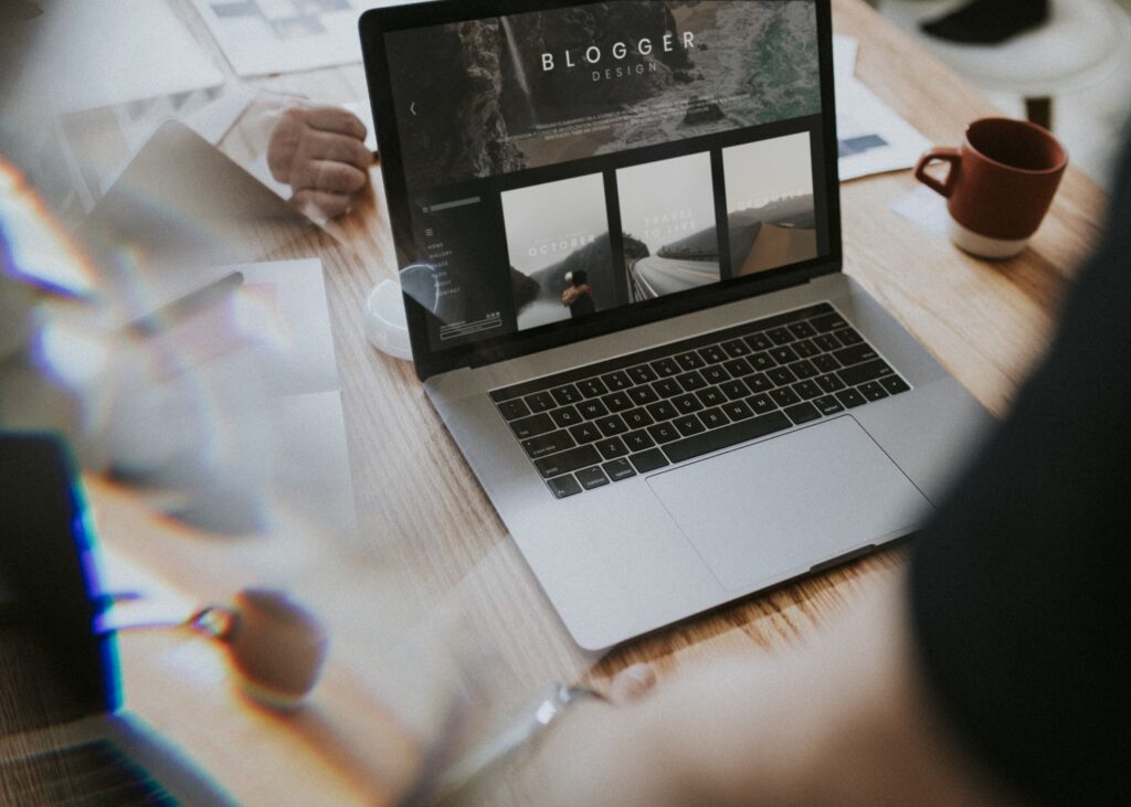 A laptop showing a blog website on its screen sits on a wooden desk alongside a red mug and papers.