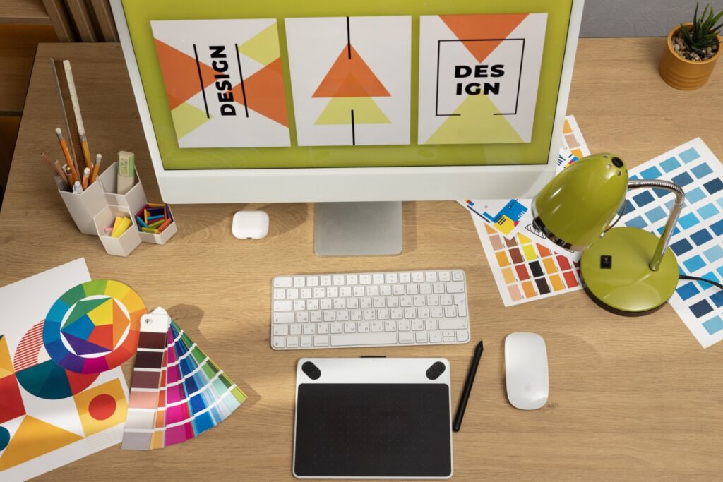Overhead shot of a creative designer's wooden desk featuring a computer monitor displaying abstract geometric designs, a white keyboard and mouse, a black drawing tablet with a stylus, color swatches, a green desk lamp, and various art supplies.