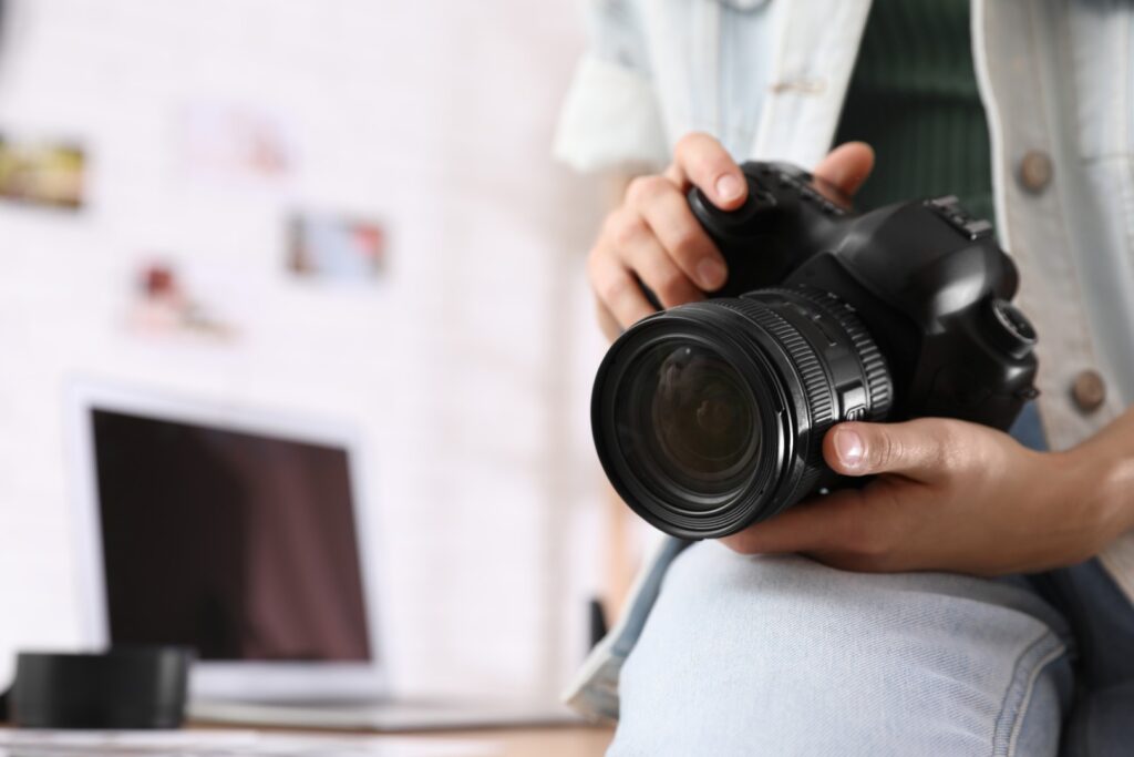 A close-up shot of a photographer holding a black DSLR camera with a large lens, with a blurred office background featuring a laptop and photos on the wall.