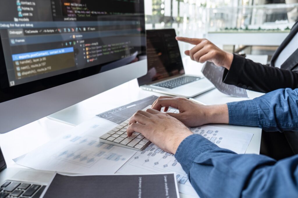 A close-up shot shows two software developers collaborating on a project in a bright office. One person types code on a computer keyboard with a large monitor displaying code, while the other points toward a laptop screen. Flowcharts and diagrams are scattered on the desk.