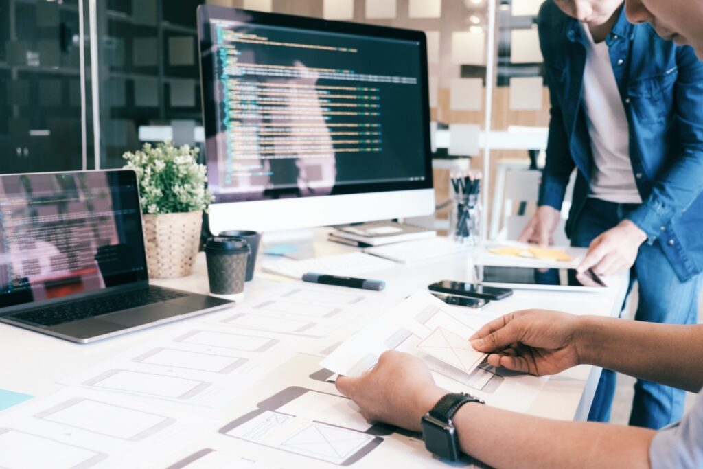 A developer holds up a paper wireframe showing mobile app designs while collaborating with colleagues in an office with code displayed on monitors.