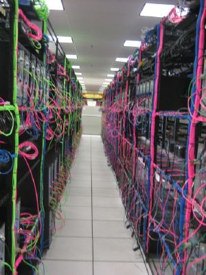 Down the aisle of a large server room, rows of black server racks with colorful cables can be seen.