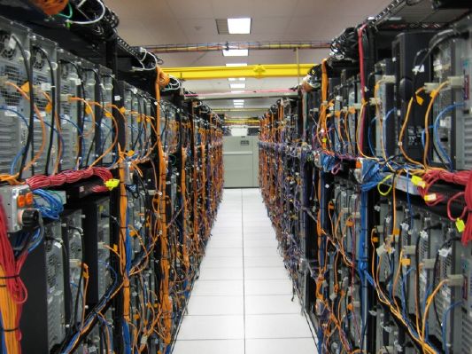 A straight, white-floored corridor runs down the middle of a data center, flanked on both sides by tall racks of servers loaded with a dense tangle of yellow, blue, and red cables.