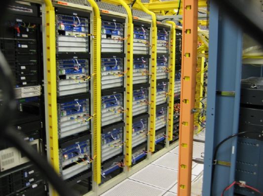 Rows of server racks with blue and white equipment and yellow overhead cable trays fill a data center.