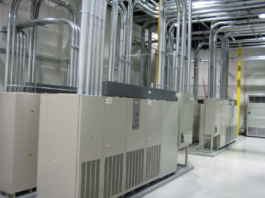 Rows of large beige server cabinets with complex overhead metal conduit and ventilation pipes in a data center.