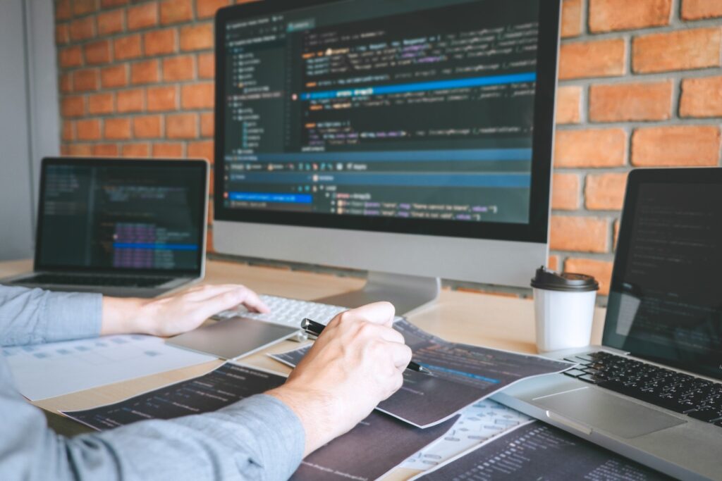 A developer writes code at a desk surrounded by multiple monitors, laptops, and printed code snippets against a brick wall.