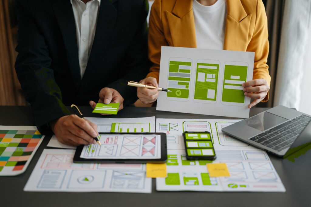 Two web designers working together at a desk, surrounded by wireframes, sketches, a tablet, and a laptop.