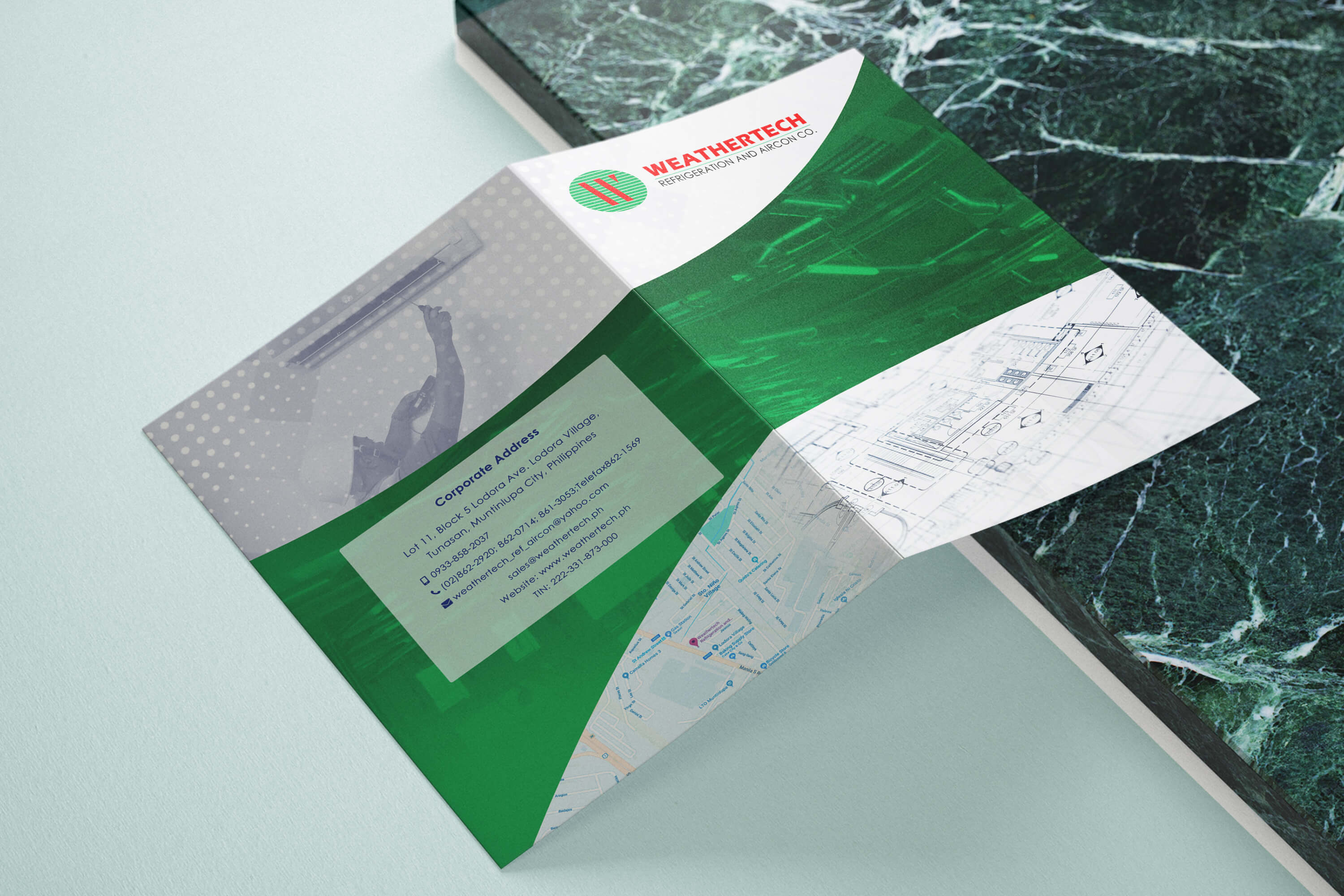 An open tri-fold brochure for Weathertech Refrigeration and Aircon Co. sits on a surface against a green marbled backdrop, displaying corporate contact info and a map.