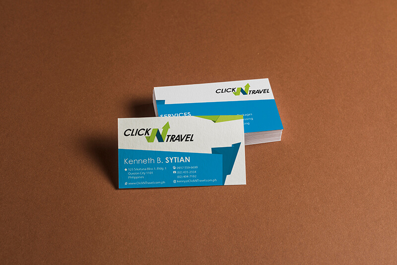 Two white business cards for Click N Travel, one upright in the foreground and one stacked in the background, rest on a brown background.