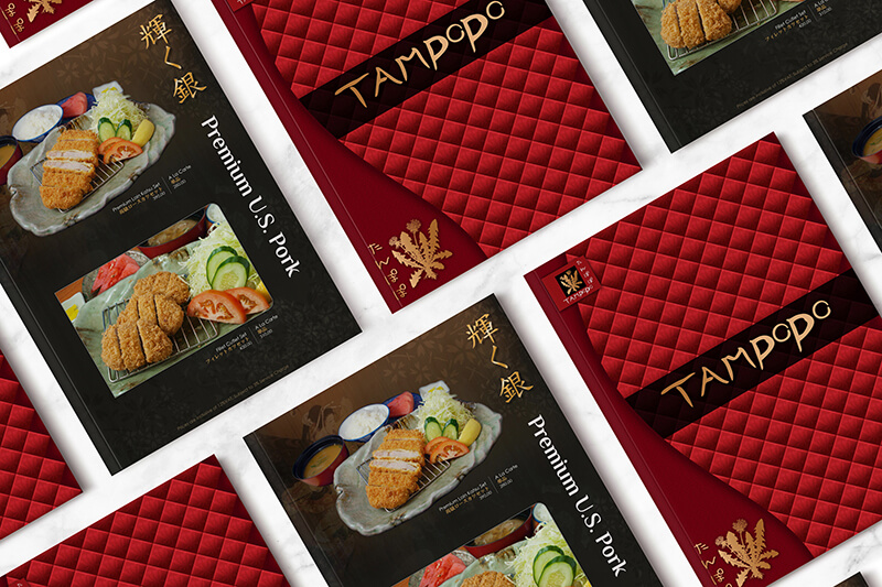 Several open menus of the Japanese restaurant Tampopo, featuring images of pork cutlets and red quilted covers, are arranged on a white marble surface.