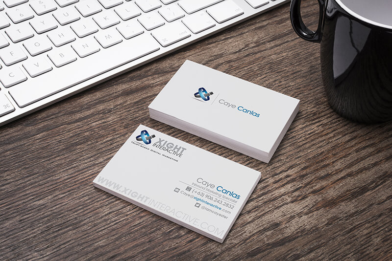A wooden desk with a white keyboard, a black mug, and two business cards featuring the Xight Interactive and Caye Canlas branding.