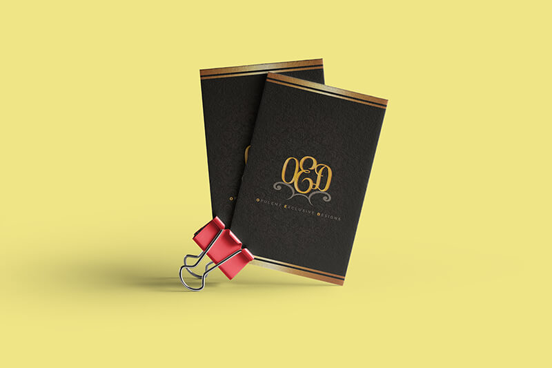 Two black and gold business cards featuring the "OED" monogram are held together by a red binder clip against a solid yellow background.