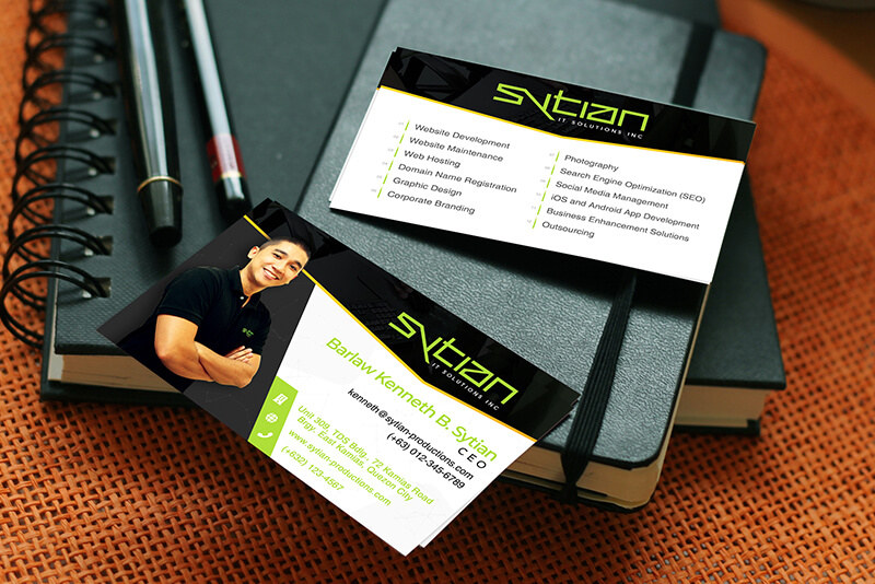 Two black and white Sytian IT Solutions business cards resting on top of a black notebook with two pens.