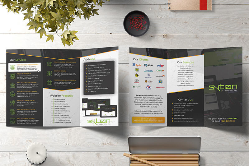 A trifold brochure for Sytian IT Solutions is laid open on a wooden table, surrounded by office supplies like a bowl of paperclips and a potted plant.