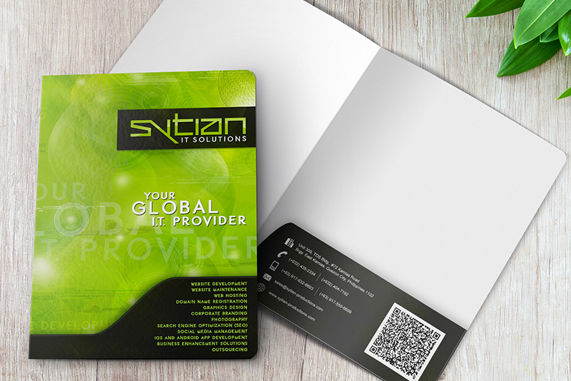 A green and black Sytian IT Solutions folder open on a wooden surface, with a list of services on the front cover and contact information on the inside pocket.