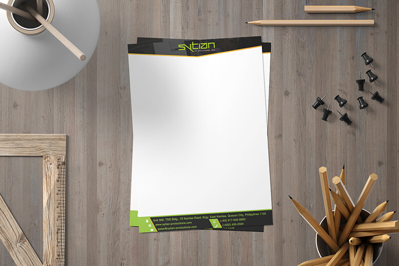 A stack of Sytian letterhead paper rests on a wooden desk alongside a ruler, pencils, and pushpins.