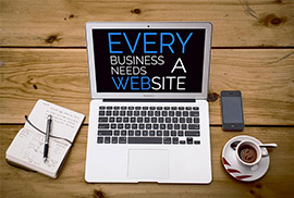 Overhead view of a silver laptop displaying the text 'Every business needs a website' on its screen, surrounded by a notebook, pen, smartphone, and cup of coffee on a wooden desk.