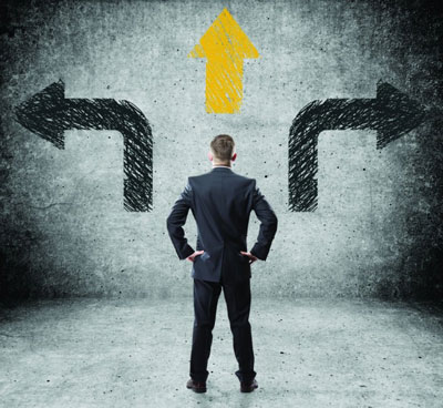 A businessman standing before a textured wall, viewing three painted directional arrows: a black arrow pointing left, a black arrow pointing right, and a bright yellow arrow pointing straight up.